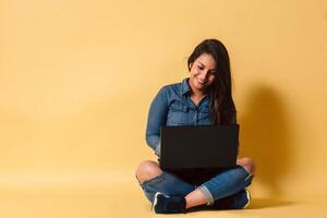 Full length of a happy latinx woman holding laptop computer sitting on floor over yellow background. photo