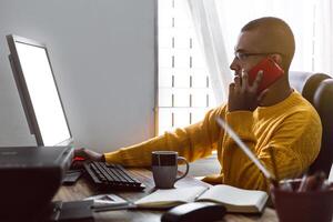 joven adulto latín americano hombre haciendo un llamada con su teléfono mientras trabajando en su computadora. foto
