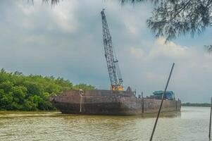 A barge with a crane on top is traveling along a river in a mangrove forest area photo