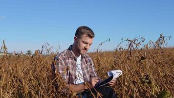 A farmer or agronomist sits in the middle of a mature soybean in a field and makes notes on a tablet with documents video