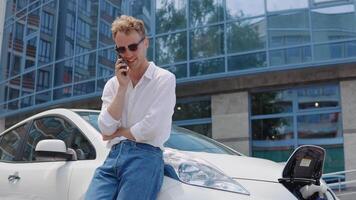 elegante moderno joven Rizado hombre en pantalones y un blanco camisa en el yarda de un residencial complejo hablando en un célula teléfono, propensión en un eléctrico coche mientras eso es cargando video
