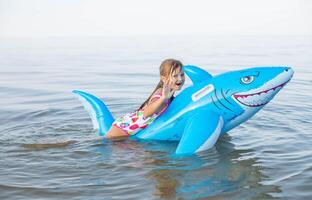 Happy girl of European appearance age of 7 swimming on an inflatable big shark toy in the sea.Family summer vocation concept. photo