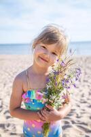 Young happy child girl of European appearance age of 6 with flowers is smiling on the beach at sunny summer day,tropical summer vocations,holidays.A child enjoys the sea.Family holidays concept.Vertical photo. photo