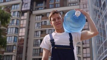 Courier holds a large can of drinking water on his shoulder against the backdrop of a modern high-rise building and looks into the camera. Water delivery home video