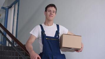 A courier in a white T-shirt and blue overalls stands on the stairs in a high-rise building holding a box in one hand video