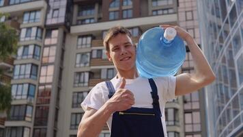 agua entrega hogar. mensajero sostiene un grande lata de Bebiendo agua en su hombro en contra el fondo de un moderno Alto edificio y mira dentro el cámara video