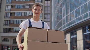 levering arbeider in een wit t-shirt en blauw overall houdt drie dozen Aan de achtergrond van modern hoogbouw gebouwen. uitdrukken levering video