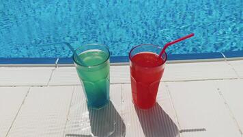 Blue and red cocktails with ice stand on side pool, against background of pool, blue water. Summer pool party, vacation video