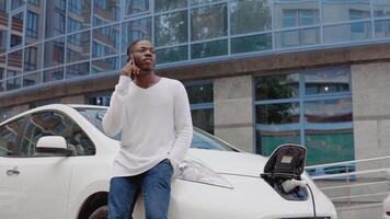 elegante africano americano hombre en el yarda de un residencial complejo hablando en un célula teléfono, propensión en un eléctrico coche mientras eso es cargando video