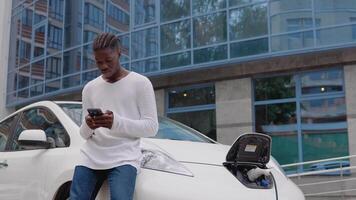 elegante joven africano americano hombre en pie cerca un eléctrico coche cargando y deslizar en su teléfono inteligente video