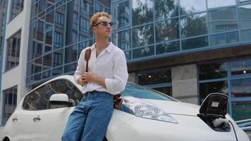 Stylish modern young curly man with a stylish city briefcase on his shoulder sits near his electric car while he charges video
