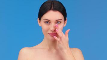 European young woman applies face cream on her cheek on blue background. Face cream advertising. Studio shot video