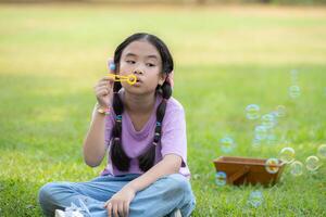 niña sentado en el parque con soplo aire burbuja, rodeado por verdor y naturaleza foto