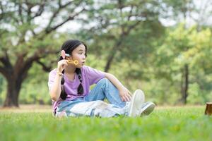 niña sentado en el parque con soplo aire burbuja, rodeado por verdor y naturaleza foto