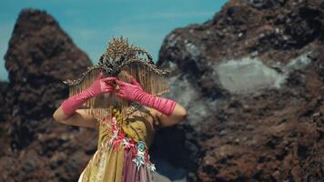 Person in ornate costume with headpiece and pink gloves posing against a rocky landscape under a clear sky. video