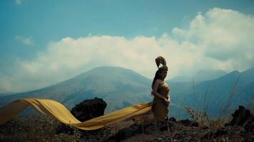 Woman in flowing dress standing on mountain with arms raised, scenic landscape background. video