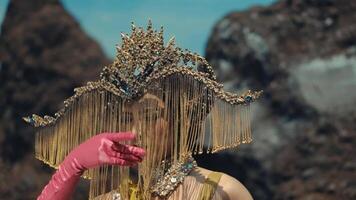 Surreal portrait of a person with an artistic headdress and pink gloves, posing against a rocky background. video