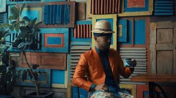Stylish man in orange jacket and hat drinking coffee against colorful wooden background. video