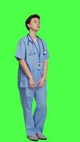 Side view Portrait of medical assistant smiling and posing with confidence, standing against greenscreen backdrop. Nurse wears blue scrubs and stethoscope, feeling successful with health expertise. Camera A. video