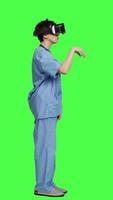 Profile Health specialist using vr headset to examine patient diagnosis, stands against greenscreen backdrop. Nurse works with artificial intelligence virtual reality glasses, three dimensional. Camera A. video