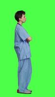 Profile Portrait of confident medical assistant posing with arms crossed in studio, standing against greenscreen backdrop. Successful nurse with healthcare expertise wearing blue scrubs uniform. Camera A. video