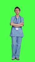 Front view Portrait of smiling medical assistant poses with arms crossed, showing confidence dressed in blue hospital scrubs. Successful nurse standing against greenscreen backdrop, health specialist. Camera A. video