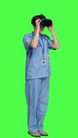 Side view Medical assistant uses interactive vr glasses to check disease treatment, looking at patient examination files with virtual reality headset and lens. Nurse against greenscreen backdrop. Camera A. video