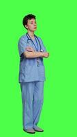 Side view Portrait of confident medical assistant posing with arms crossed in studio, standing against greenscreen backdrop. Successful nurse with healthcare expertise wearing blue scrubs uniform. Camera A. video