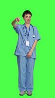 Front view Displeased medical assistant shows thumbs down in studio, presenting her disapproval and disagreement with dislike symbol against greenscreen. Nurse feeling unhappy with rejection sign. Camera A. video