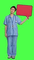 Front view Medical assistant holding a red speech bubble icon against greenscreen, creating advertisement with empty copyspace billboard sign. Nurse shows cardboard poster, marketing announcement. Camera A. video