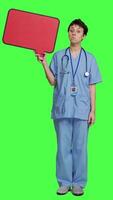 Front view Medical assistant holding a red speech bubble icon against greenscreen, creating advertisement with empty copyspace billboard sign. Nurse shows cardboard poster, marketing announcement. Camera A. video