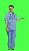 Front view Medical assistant pointing at something aside and doing web commercial, creating advertisement against greenscreen backdrop. Young nurse with scrubs showing a thing to left or right sides. Camera A. video