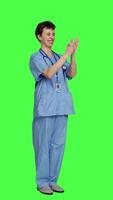 Side view Smiling medical assistant applauding someone in studio, celebrating success and cheering against greenscreen backdrop. Young nurse clapping hands and feeling happy about achievement. Camera A. video