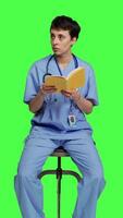 främre se ung sjuksköterska studerar för de läkare examen förbi läsning bok i studio, Sammanträde på stol mot grönskärm. specialist åtnjuter sjukvård industri litteratur, framställning till bli en läkare. kamera a. video