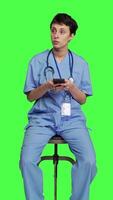 Front view Nurse browsing online websites on smartphone app in studio, sitting on a chair against greenscreen backdrop. Medical specialist in scrubs using mobile phone and social media texting. Camera A. video