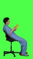 Profile Health specialist is reading a literature book to acquire physician specialization and surgeon expertise, sitting in a chair. Nurse studying healthcare industry lecture against greenscreen. Camera A. video