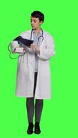 Front view Healthcare specialist using a tonometer to measure high blood pressure and pulse, presenting tool used at checkup examinations. Doctor in white coat stands against greenscreen backdrop. Camera A. video