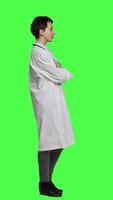 Profile Portrait of successful physician standing with arms crossed against greenscreen backdrop, wearing a white coat and stethoscope. Doctor feeling confident with healthcare expertise. Camera A. video