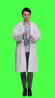 Front view General practitioner with a white coat using stethoscope in studio, preparing to examine patients at checkup appointments. Female doctor in uniform standing against greenscreen backdrop. Camera A. video