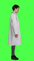 Profile Physician saying no and showing negative reaction symbol in studio, wearing a white coat and standing against greenscreen backdrop. Woman medic feeling displeased, rejection sign. Camera A. video