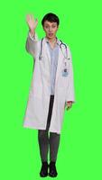 Front view Woman medic waving hello and asking people to come over at checkup, inviting patient to come closer and greets them. Skilled physician with white coat and stethoscope against greenscreen. Camera A. video