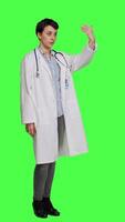 Side view Woman medic waving hello and asking people to come over at checkup, inviting patient to come closer and greets them. Skilled physician with white coat and stethoscope against greenscreen. Camera A. video