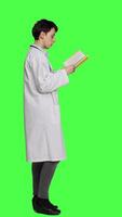 Profile Medic specialist reading a healthcare literature book to develop her medical expertise in the industry, stands against greenscreen backdrop. Clever doctor in white coat enjoys lecture. Camera A. video