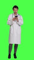 främre se läkare navigerande på social media appar till text människor, står mot grönskärm bakgrund. läkare i vit täcka bläddring webbplatser uppkopplad, framställning kolla upp möten. kamera a. video