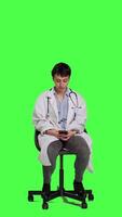 Front view General practitioner texting messages on mobile phone, killing time while she is waiting for patients to arrive at checkup. Woman physician sitting on a chair against greenscreen backdrop. Camera A. video