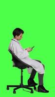 Profile General practitioner texting messages on mobile phone, killing time while she is waiting for patients to arrive at checkup. Woman physician sitting on a chair against greenscreen backdrop. Camera A. video