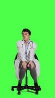 Front view Physician in white coat waiting for patients at consultations, feeling impatient sitting on a chair against greenscreen backdrop. Medic practitioner with stethoscope waits for people. Camera A. video