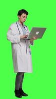 Side view Confident physician searching for new medicaments to treat disease, using laptop to browse on online web pages against greenscreen backdrop. Medic in white coat checks internet sites. Camera A. video