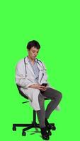 Side view General practitioner texting messages on mobile phone, killing time while she is waiting for patients to arrive at checkup. Woman physician sitting on a chair against greenscreen backdrop. Camera A. video