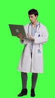 Front view Confident physician searching for new medicaments to treat disease, using laptop to browse on online web pages against greenscreen backdrop. Medic in white coat checks internet sites. Camera A. video